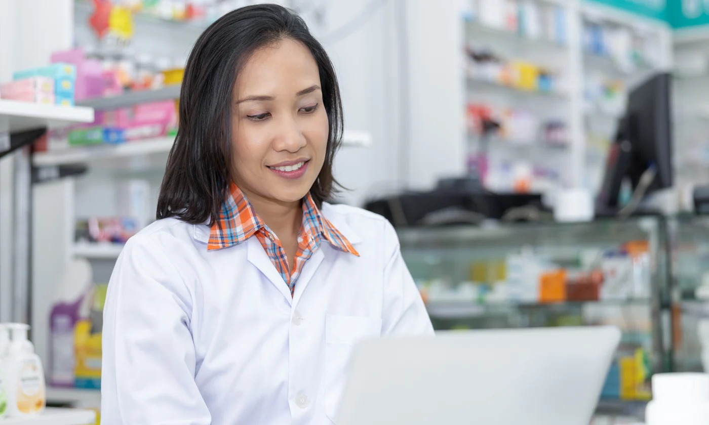Female pharmacist sitting at desk in pharmacy entering data on laptop